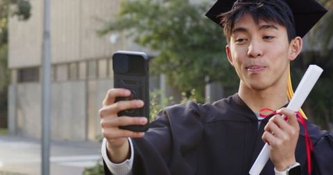 Asian college graduate taking selfie in cap and gown holding diploma on campus courtyard