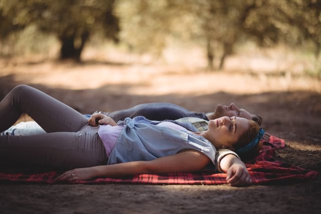 Young couple lying on a blanket in an olive farm, enjoying a peaceful moment together. Ideal for use in advertisements for romantic getaways, nature retreats, or lifestyle blogs focusing on outdoor activities and relationships. Can also be used in articles about relaxation, rural living, and couple bonding experiences.
