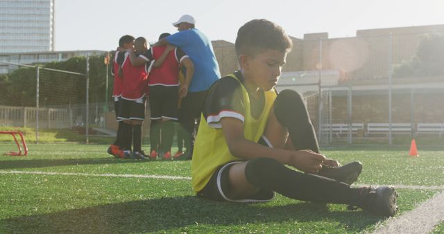 This poignant scene shows a youthful boy sitting alone on a soccer field, feeling dejected while his teammates huddle together in the background. Perfect for visuals highlighting themes of teamwork, inclusion, youth sports, or feelings of isolation. Useful for promotional materials for sports organizations, campaigns on anti-bullying or loneliness, or illustrating children's diverse emotional experiences.