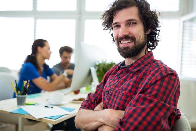 A young male graphic designer is smiling with arms crossed in a modern office. He is dressed in casual attire, indicating a relaxed and creative environment. In the background, colleagues are seen collaborating, adding to the bustling office atmosphere. This image is suitable for illustrating concepts like creativity, teamwork, modern workspaces, and professional life in marketing materials, websites, and blogs.