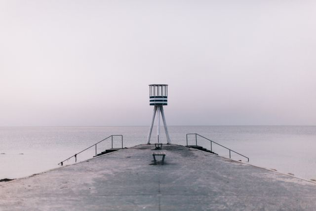A minimalist scene with an empty pier extending to the ocean and a solitary lifeguard tower at the end. The calm waters and serene horizon underscore a feeling of peacefulness and solitude. Ideal for use in projects focusing on themes of tranquility, minimalism, and coastal environments. Suitable for backgrounds, travel blogs, mindfulness apps, or motivational materials emphasizing serenity and contemplation.