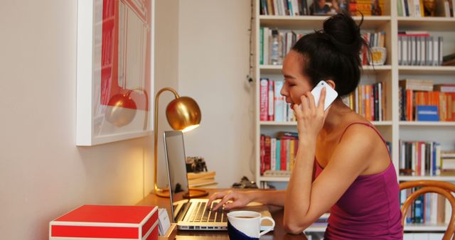 Young woman multitasking at home office, speaking on phone while working on laptop. Ideal for articles on remote work, freelance lifestyle, and modern workspaces. Depicts productivity, professional work atmosphere, and home office setup.