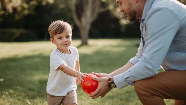 Father passing red ball to smiling little boy on sunny lawn for family playtime