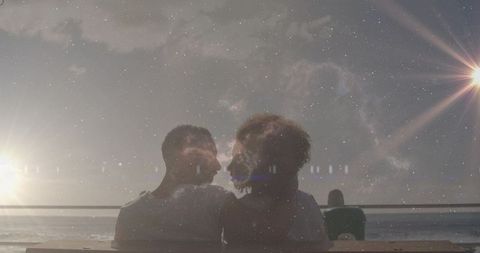 Couple leaning on bench watching sunset over ocean with starry sky double exposure