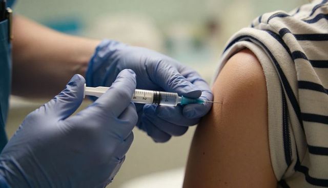 Clinician administering vaccine into upper arm wearing blue gloves patient in striped shirt