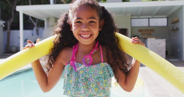 A young girl with curly hair, wearing swimwear and a pair of goggles around her neck, playfully holding yellow pool noodles. She is enjoying time by the poolside under the bright sun, symbolizing fun and carefree summer days. Ideal for use in advertisements for pool products, summer camps, vacation packages, and family activities.