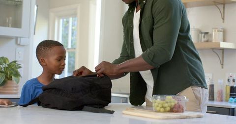 African american father packing school backpack with son on kitchen island in morning