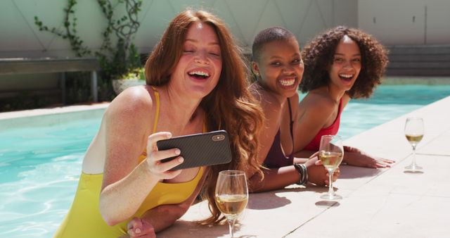 Three diverse women are laughing and enjoying each other’s company by a pool during a summer day. They are leaning on the pool edge, taking a selfie with a smartphone. They have drinks placed on the edge, suggesting a relaxed, social gathering. This type of scene is perfect for promoting summer activities, friendship, leisure, lifestyle brands, or travel advertising.
