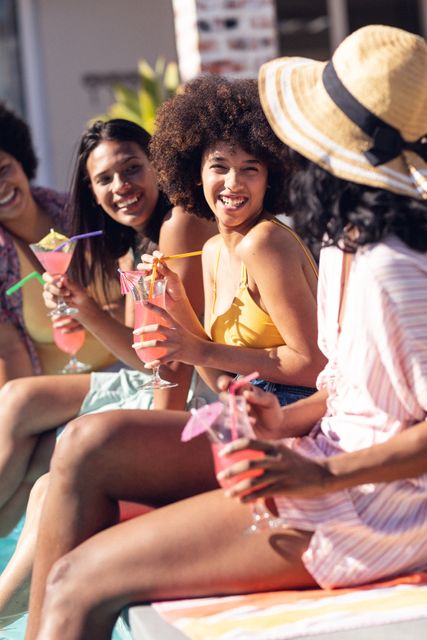 Cheerful biracial female friends having cocktails while chatting at poolside in summer party. Drink, happy, unaltered, friendship, togetherness, social gathering, enjoyment and weekend activities.