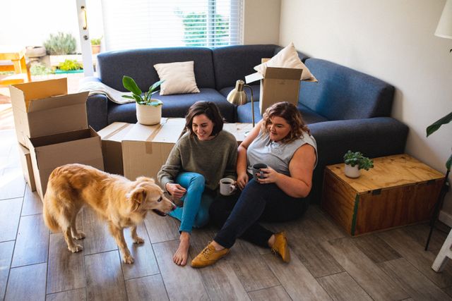 Lesbian couple sitting on floor with dog, drinking coffee and enjoying new home. Ideal for themes of moving, new beginnings, domestic life, LGBTQ representation, and home decor.