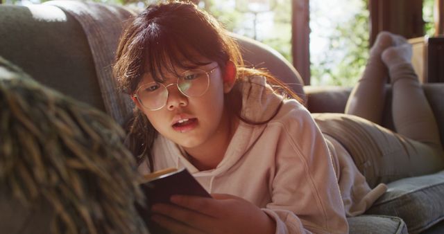 Asian girl smiling while reading a book while siting on the couch at home. childhood, hobby and leisure activity concept