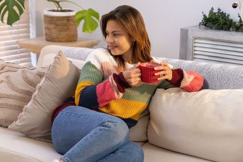 Woman sitting on sofa holding red mug wearing multicolor chunky knit sweater enjoying cozy moment