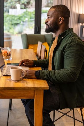 Working remotely african american man using laptop at cozy wooden table with coffee mug