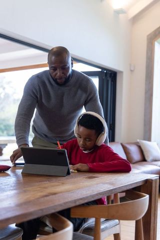 African american father guiding son doing homework on tablet in light-filled home workspace
