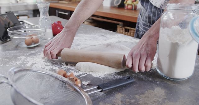 A baker is rolling out dough using a rolling pin on a floured counter. Surrounding the dough are kitchen utensils such as a strainer, a jar of flour, eggs, and other baking ingredients. This image is useful for websites and publications related to baking, cooking tutorials, homemade food preparation, or any culinary-related content. It emphasizes hands-on cooking and the baking process.