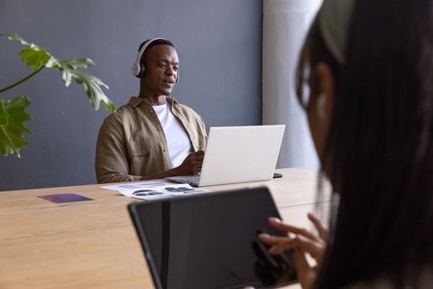 Diverse coworkers collaborating at office table with laptop, tablet, stylus and headphones