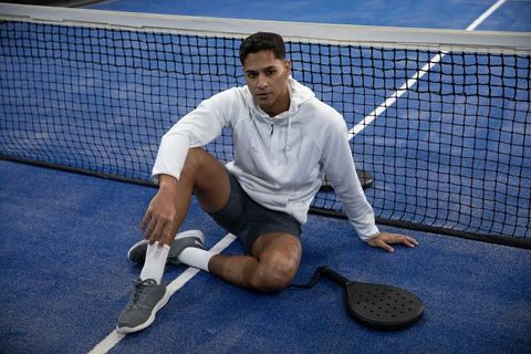 Padel player resting by net on blue court wearing white hoodie, shorts and knee tape