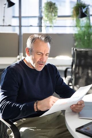 Middle-aged man reviewing documents at sunlit modern office table, pointing at paper