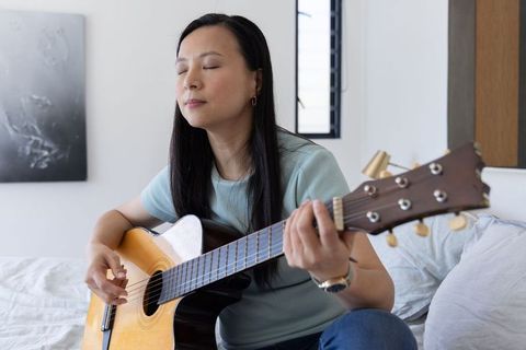 Asian woman practicing acoustic guitar on bed in minimalist bedroom, relaxing and focused