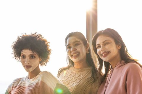 Multicultural women wearing facial masks, smiling and relaxing by sunlit window, promoting wellness