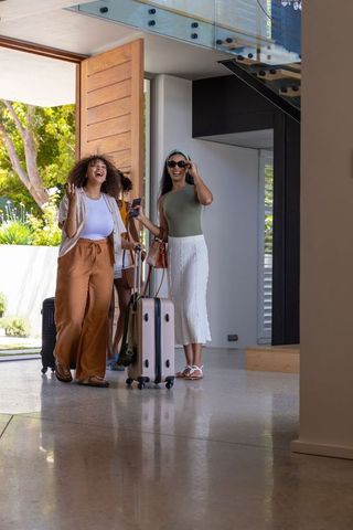 Multicultural friends entering modern home with rolling luggage through wooden pivot door