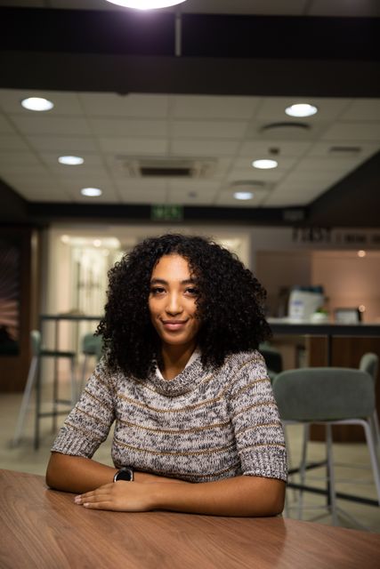 This image depicts a confident biracial professional woman working late in a modern office. She is sitting by a desk, looking at the camera and smiling. Ideal for use in business, career, and workplace-related content, highlighting themes of dedication, productivity, and modern work environments.