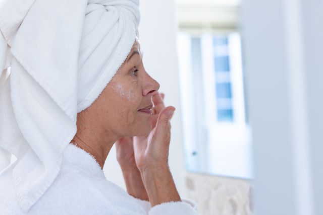 This image depicts a mature woman with a towel wrapped around her head, applying moisturizer to her face while looking in the bathroom mirror. Ideal for use in articles or advertisements related to skincare, beauty routines, senior health, and wellness. It can also be used in promotional materials for skincare products targeting older demographics.
