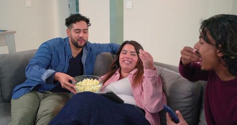 Diverse friends sharing popcorn during movie night on cozy gray couch with tablet