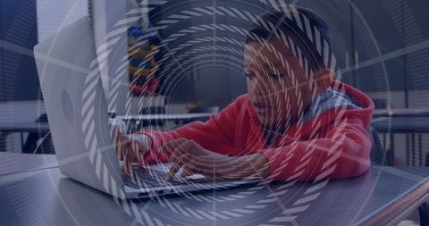 Preteen boy typing on laptop wearing red hoodie and knit beanie in school cafeteria