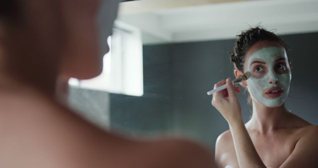 Woman applying facial mask in front of bathroom mirror, reflecting a moment of self-care and relaxation. Useful for skincare product advertisements, wellness and beauty blogs, and DIY self-care tutorials.