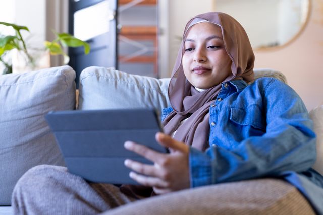Smiling biracial woman in hijab using tablet relaxing on couch at home, copy space. Happiness, communication, relaxation, inclusivity and domestic life.