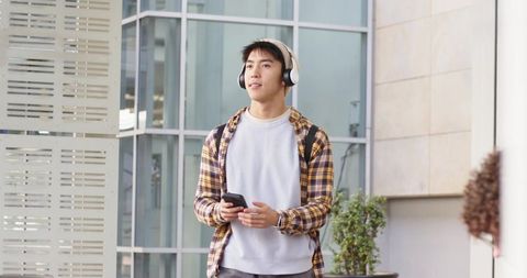 Asian man walking with headphones and smartphone by modern office building, casual commuter lifestyl