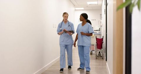 Multiracial female nurses walking hospital corridor reviewing tablet and discussing care