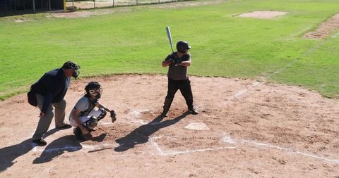 Batter Batting at Home Plate, Catcher Squatting and Umpire Observing Sunlit Baseball Field