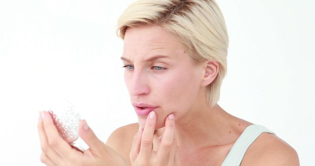 Blonde woman holding a compact mirror and touching chin, highlighting skincare and beauty routines. Ideal for use in health and beauty promotions, skincare product ads, tutorials on facial care, and wellness blogs.