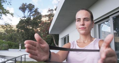 Focused woman stretching resistance band on balcony wearing pink top and fitness tracker