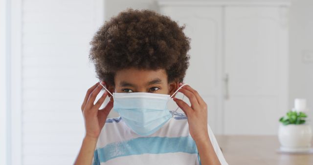 Portrait of an African American boy with afro hair putting a face mask on, looking to the camera. Social distancing and self isolating at home during Coronavirus Covid 19 quarantine lockdown.