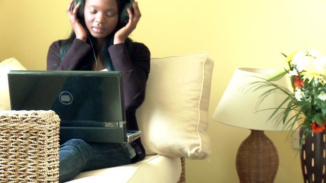 Young woman in casual attire sitting on comfortable sofa, wearing headphones and using laptop. Ideal for illustrating leisure time, technology use at home, or relaxation activities. Perfect for lifestyle blogs, articles on work-life balance, or digital music streaming.