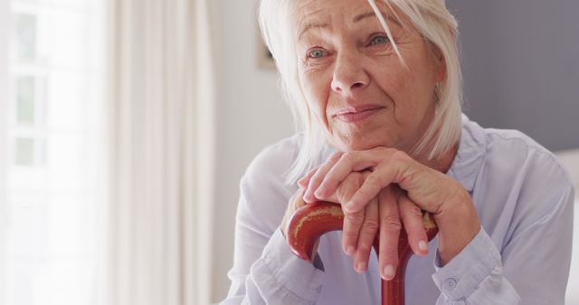 Elderly woman holding a walking stick, sitting indoors, looking content and relaxed. Ideal for use in health and wellness, retirement planning, elderly care services, or home and family focused publications.