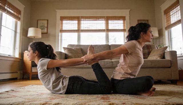 Mother and daughter practicing partner yoga on living room rug holding bow pose at morning