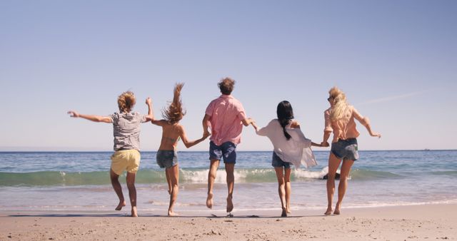 Group of friends holding hands and jumping into ocean water from sandy beach. Perfect for campaigns promoting summer vacations, friendship, youth lifestyle, and outdoor activities.