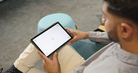 Indian man sitting on teal sofa holding tablet with blank screen for mockup