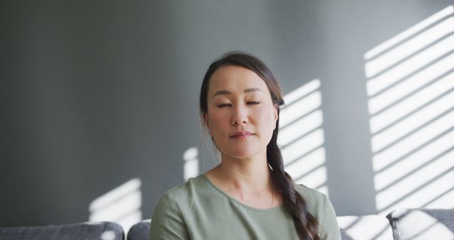Woman sitting on couch with eyes closed, meditating near window with light filtering through blinds. Useful for illustrating mindfulness, relaxation techniques, mental health awareness, or demonstrating indoor activities focused on reducing stress.