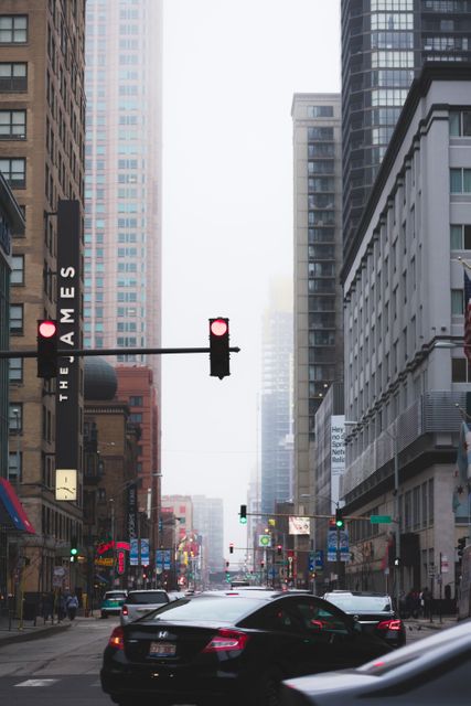 Foggy city street crowded with modern high-rise buildings and urban traffic. Red traffic light dominating the scene with cars waiting and various street signs. This dynamic image can be used for conveying messages about city life, urban development, traffic control, and city planning. Ideal for urban studies, real estate promotion, and transportation services.