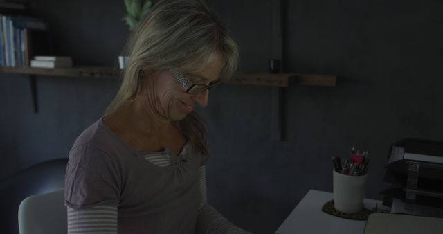 Smiling mature woman working at desk in dimly lit home office - Download Free Stock Images Pikwizard.com