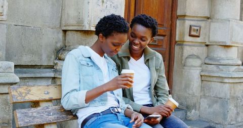 Smiling African American Twin Sisters Sharing Coffee and Smartphone in Urban Setting