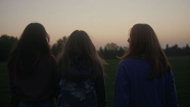 Three girls standing and watching dusk sky over meadow at sunset, silhouetted trio