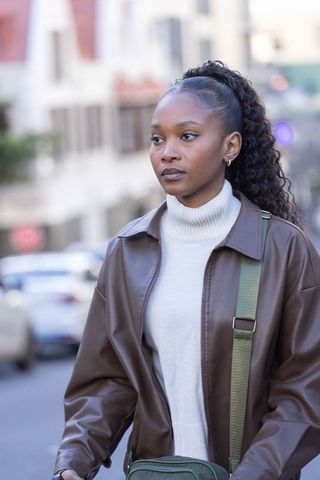 Young woman walking in brown leather jacket and white turtleneck with green crossbody bag