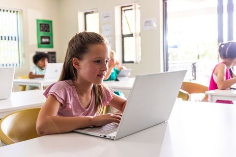 Elementary girl using laptop in modern classroom with diverse students learning and working