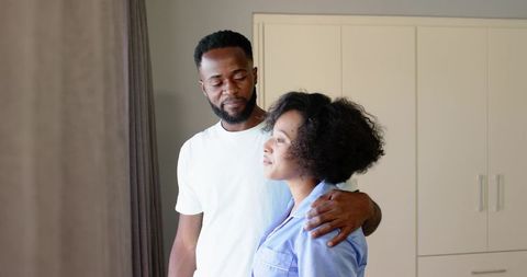 African American Couple Embracing by Window in Bedroom, Man Holding Partner Affectionately